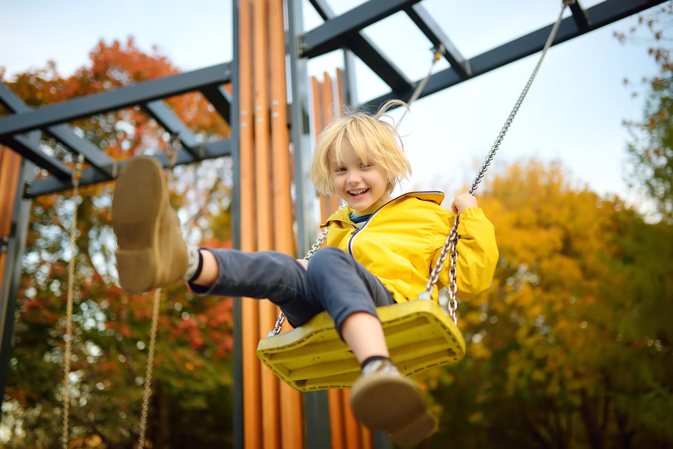 A child with a bright yellow raincoat and equally bright blond hair smiles while playing on a swing set.