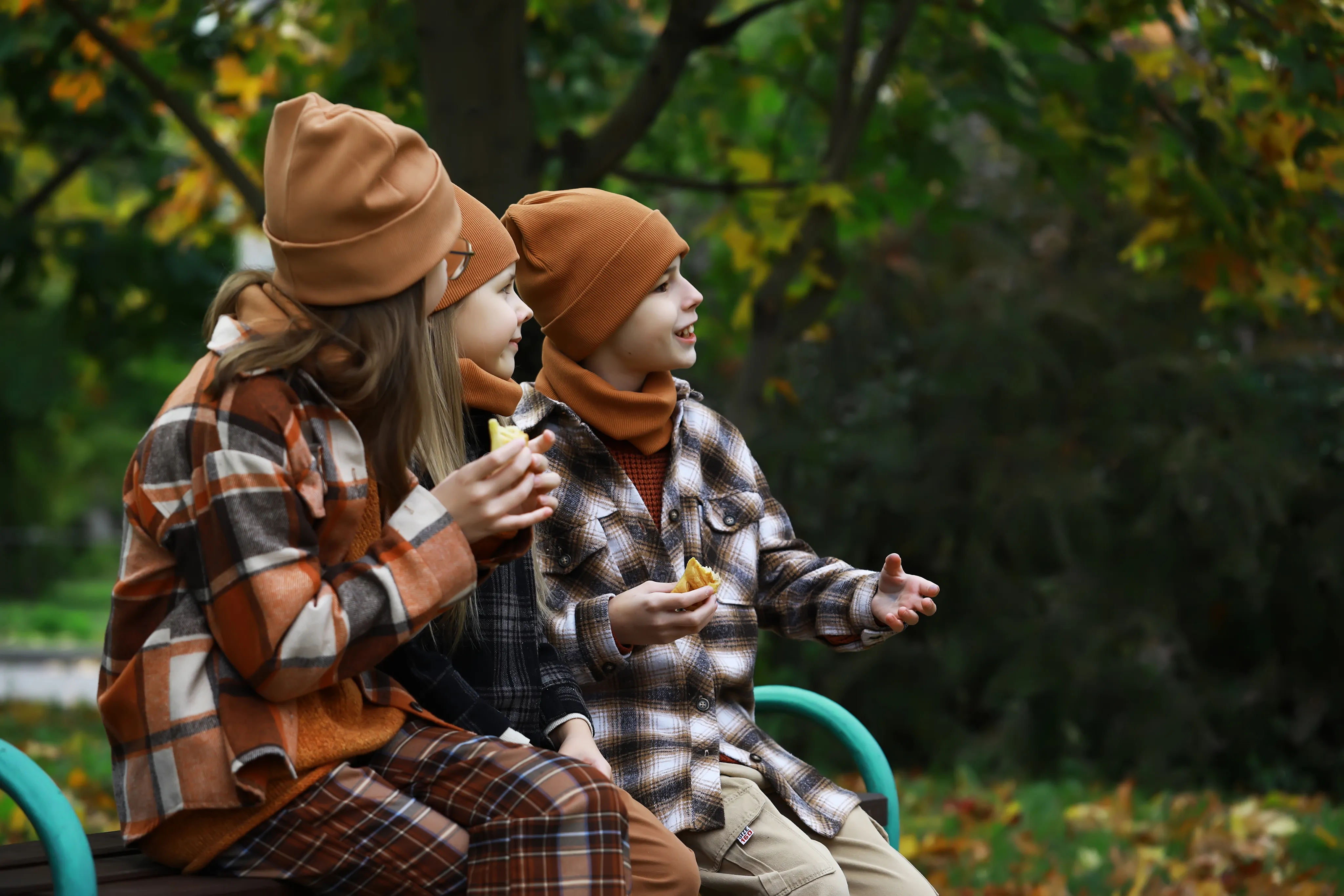 Three children, dressed in their Fall best, are seen in profile eating a snack and laughing as they look on.