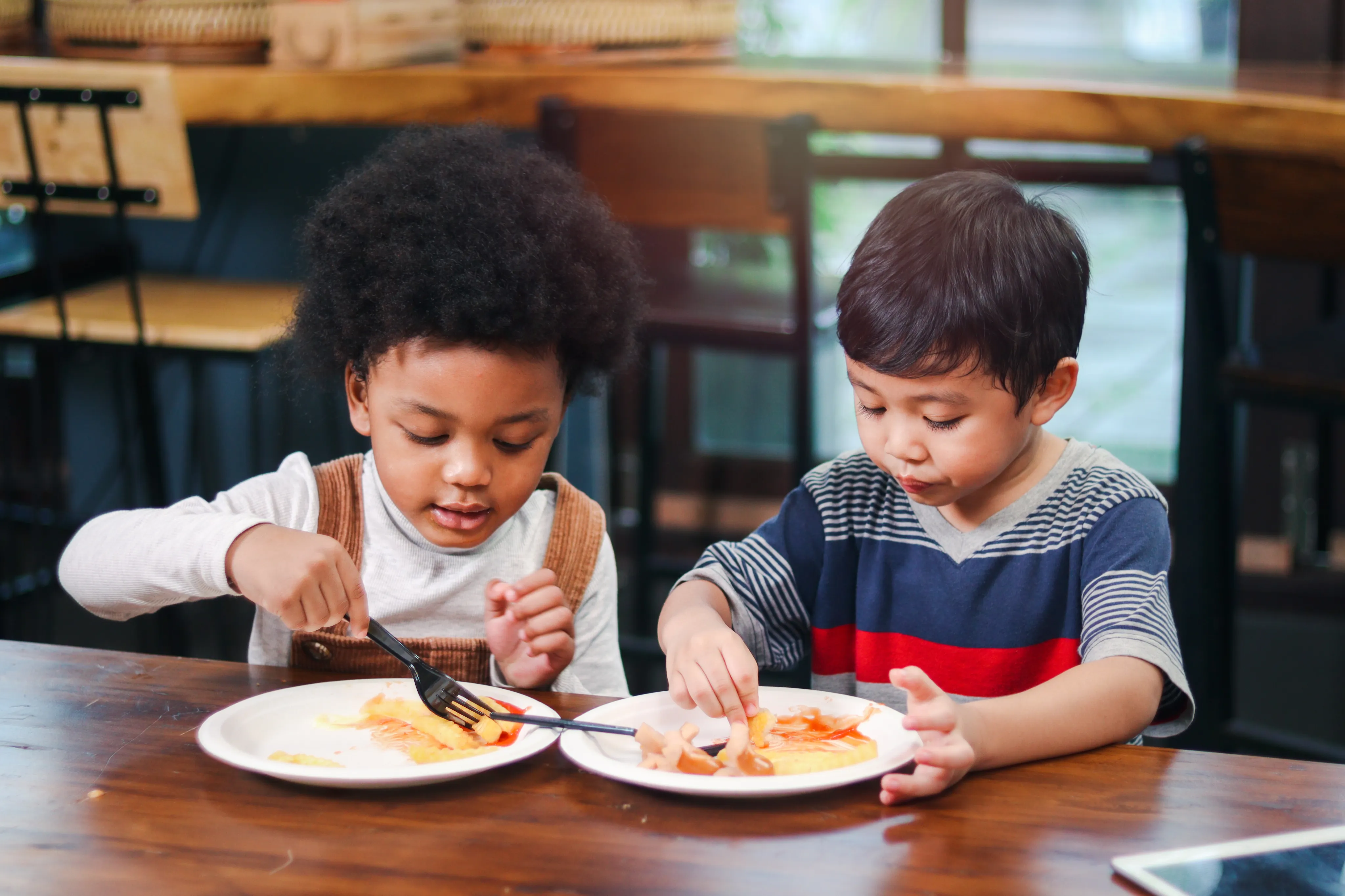 Two children sit at a table together enthusiastically enjoying their meal.